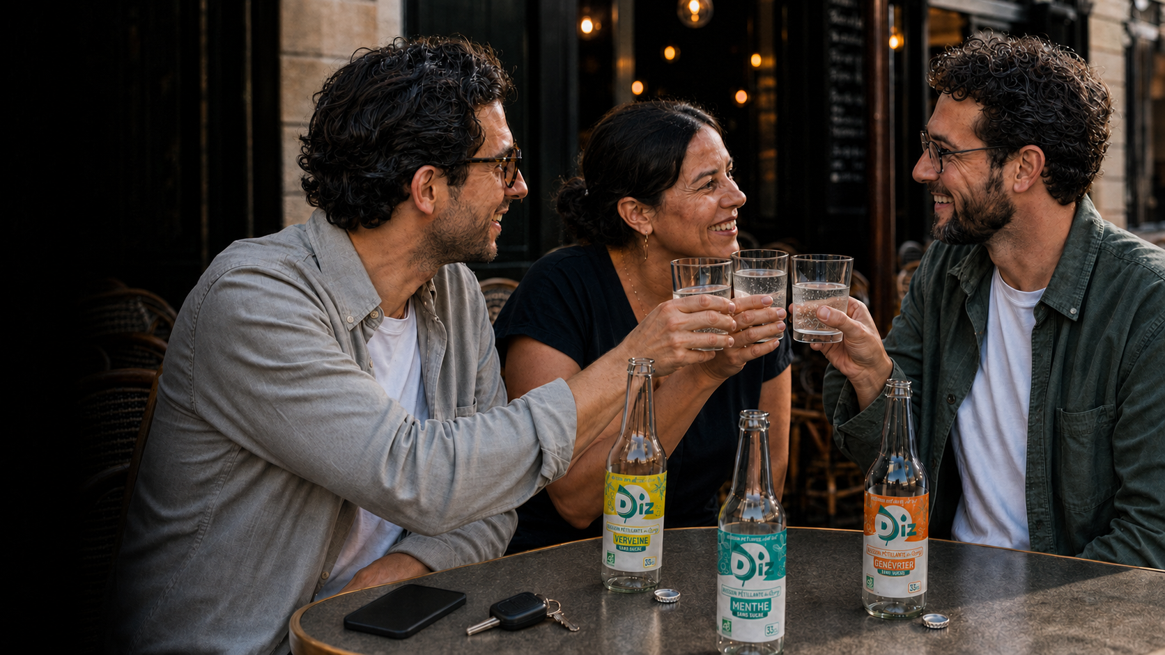 Trois amis trinquent au verre de DIZ à la terrasse d’un café, avec les bouteilles Verveine, Menthe et Genévrier sur la table